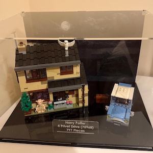 May include: A clear acrylic display case with a black base, showcasing a Lego Harry Potter set of 4 Privet Drive (75968) with 797 pieces. The set includes a detailed house, a flying owl, and a blue car.