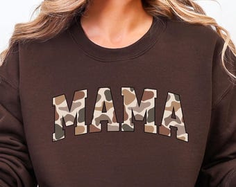 May include: A brown sweatshirt with the word "MAMA" in a camouflage pattern across the chest. The sweatshirt has a crew neck and long sleeves. The person in the image is wearing blue jeans and holding a mug with a grid pattern.