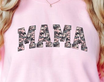 May include: A light pink sweatshirt with the word "MAMA" in a camouflage pattern across the chest. The sweatshirt has long sleeves and a crew neck. A red tumbler with a Christmas tree design is held in the right hand.