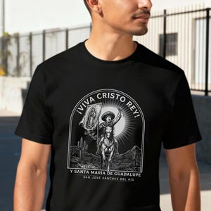 Pode incluir: Camiseta preta com um gráfico monocromático de uma pessoa a cavalo com uma bandeira, em um cenário desértico. O texto "¡VIVA CRISTO REY!" está acima da imagem, com mais texto abaixo. A camiseta é feita de um material macio.