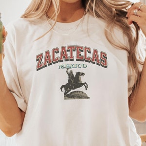May include: A white t-shirt with the text "Zacatecas Mexico" in red and blue lettering. The shirt features a vintage graphic of a person riding a horse.