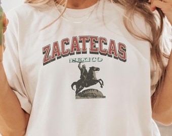 May include: A white t-shirt with the text "Zacatecas Mexico" in red and blue lettering. The shirt features a vintage graphic of a person riding a horse.