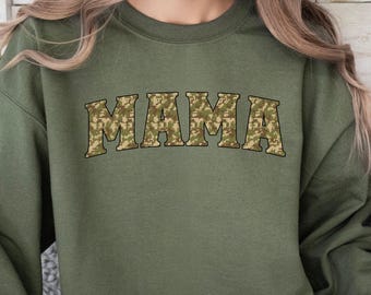 May include: Olive green sweatshirt with the word "MAMA" in a camouflage pattern across the chest. The sweatshirt has long sleeves and a crew neck. The text is in a bold, block letter style.