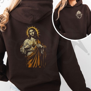 May include: A dark brown hoodie featuring a detailed graphic of Jesus with a golden halo and a heart on the back. The front of the hoodie has a smaller, matching design. The hoodie has a hood and a front pocket.