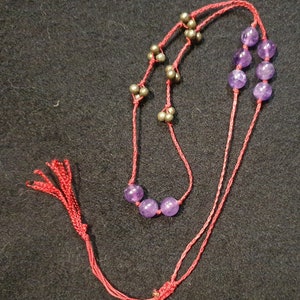 Può includere: Una collana di corda rossa con perline viola e dettagli dorati. La collana ha un fiocco all'estremità.
