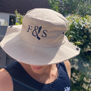 May include: A beige bucket hat with a wide brim. The hat has the embroidered letters "F & S" in navy blue. The hat has a snap on each side and is made of a light-coloured fabric, suitable for sun protection.