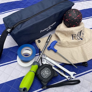 May include: A collection of tools and accessories, including a navy blue zippered pouch with "Feather & Square" text, a beige bucket hat with "F&S" embroidery, and a black and red massage ball. Other items include a tape measure, spanners, and a screwdriver.
