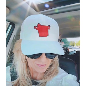 Puede incluir: Gorra de béisbol blanca con un logotipo rojo estilizado de Razorback. La gorra la lleva una persona con gafas de sol y aros. La imagen está tomada dentro de un coche con el sol brillando a través del techo corredizo.