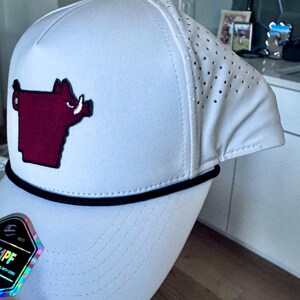May include: White baseball cap with a maroon hog logo on the front. The cap has a black rope detail and a perforated side panel. The brim is curved, and the cap has a button on top.