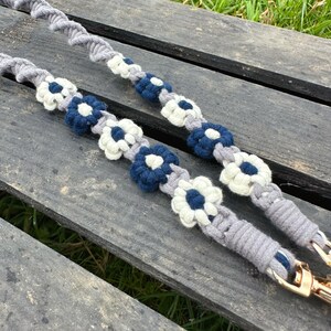 May include: Two gray macrame straps with white and navy blue flower accents. The straps have gold-colored clasps at the ends. The straps are lying on a weathered wooden surface.