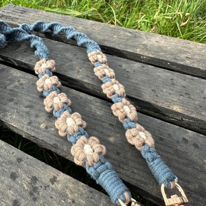 Puede incluir: Una correa de macramé azul con un diseño en espiral y detalles florales. Las flores son beige con centros blancos. La correa tiene cierres metálicos dorados.