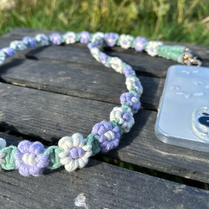 May include: A handmade phone strap featuring a floral design. The strap is made of crocheted yarn in shades of lavender, white, and green. The strap is connected to a clear phone case with a gold-colored clasp. The phone case is on a wooden surface.