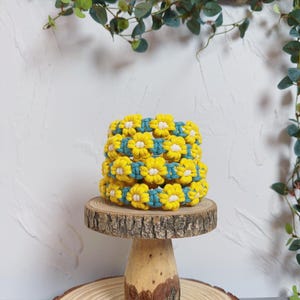 May include: A stack of three yellow and white flower-shaped macrame bracelets with teal accents. The bracelets are displayed on a small, round wooden stand with a natural edge. Green foliage is in the background.