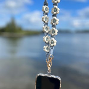 May include: A light blue macrame phone lanyard with white and light blue flower accents. The lanyard is attached to a gold-colored clasp and a clear phone case. The background is a blurred view of a lake and sky.