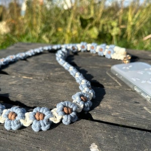 May include: A blue and white phone strap with a flower design. The strap is made of woven string and features brown centers in each flower.