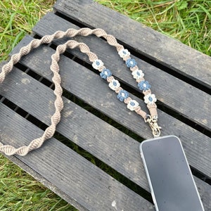 May include: A phone strap with a brown braided cord and white and blue flower accents. The strap is attached to a black phone case.