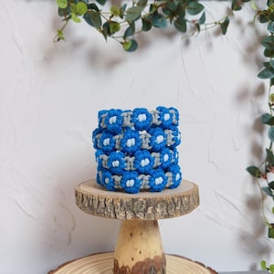 May include: A stack of blue and gray macrame bracelets with a flower design. The bracelets are displayed on a small, round wooden stand with a natural edge. Green foliage is in the background.