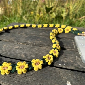 May include: A green and yellow crocheted phone strap with sunflower designs. The strap is lying on a wooden surface.