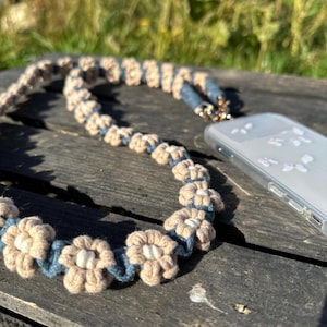 May include: A phone strap made of woven beige and blue fabric with a flower pattern. The strap is attached to a clear phone case.