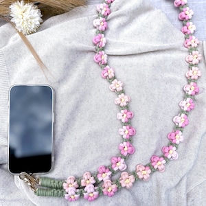 May include: A phone strap made of green and pink yarn with a flower design. The strap is made of two strands of yarn that are knotted together to form flowers. The flowers are pink and white with green centers.