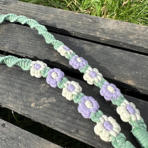 May include: Two handmade crochet straps with a floral design. The straps are light green with white, lavender, and green flower accents. The straps are lying on a weathered wooden surface.