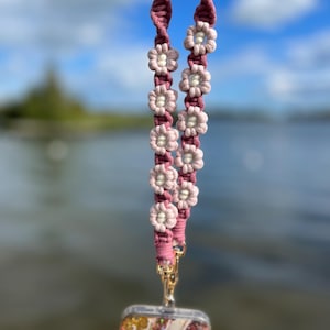 May include: A pink macrame lanyard with a floral design. The lanyard features a series of small, light pink and white flowers. A gold-colored clasp connects the lanyard to a phone case with a colorful, abstract design.