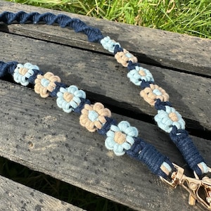 May include: A blue and beige macrame cord strap with a flower design. The strap has a gold metal clasp.
