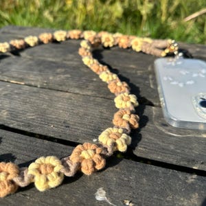 May include: A phone strap made of brown and yellow yarn, woven into a flower pattern. The strap is attached to a clear phone case.