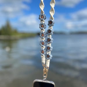 May include: Cream-colored macrame phone lanyard with blue and brown flower accents. The lanyard is attached to a clear phone case with a gold-colored clasp. The background features a blurred view of water and a blue sky.
