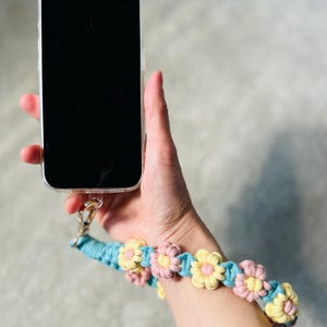 May include: A smartphone with a clear case attached to a wrist strap made of braided teal cord featuring alternating pink and yellow crocheted flower motifs. The strap connects to the phone case via a gold-toned metal lobster clasp hardware.