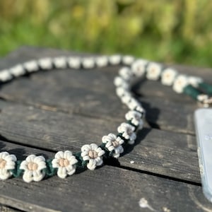 May include: A white and green phone strap with a daisy design. The strap is made of woven string and has a simple, minimalist style.