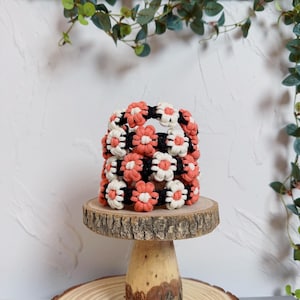 May include: A handmade, square-shaped macrame basket featuring a floral design in coral, black, and white. The basket sits on a small, round wooden stand with a natural wood grain finish. The background is a white wall with green foliage.