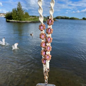 May include: A macrame lanyard with a cream-colored twisted cord and brown and lavender flower accents. The lanyard is attached to a clear phone case with a colorful glitter design and a gold-colored clasp. The background shows a lake and blue sky.