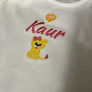 May include: White baby onesie with pink embroidery of the name "Kaur" and a small yellow lion with a pink bow. A small yellow symbol is above the name.