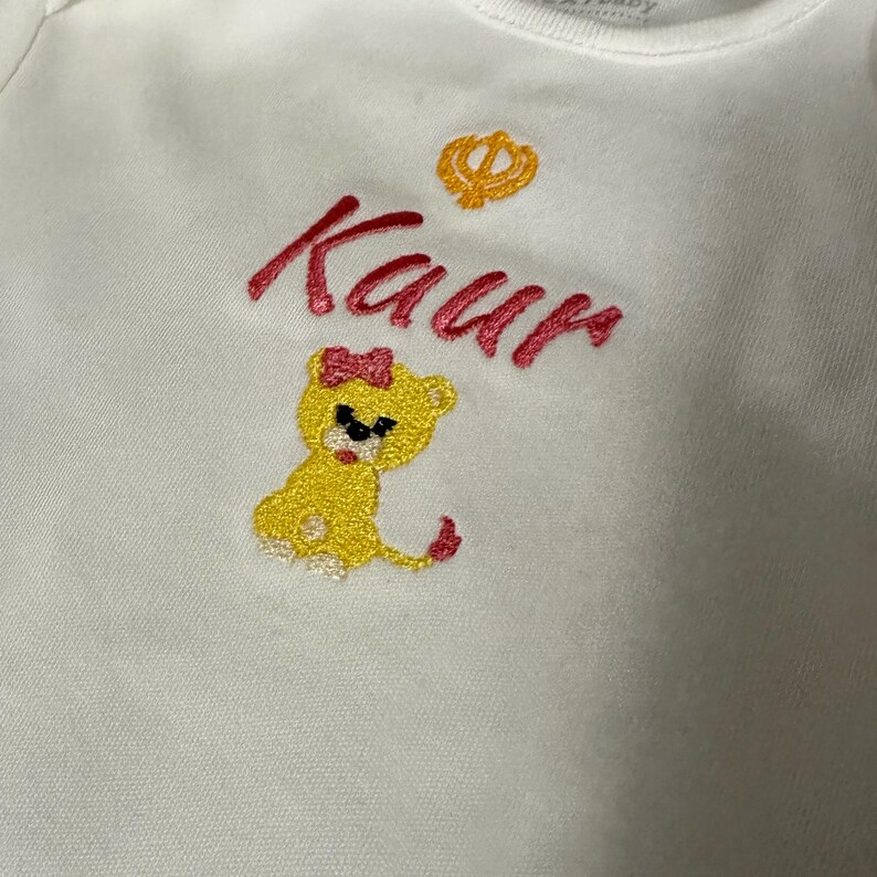 May include: White baby onesie with pink embroidery of the name "Kaur" and a small yellow lion with a pink bow. A small yellow symbol is above the name.