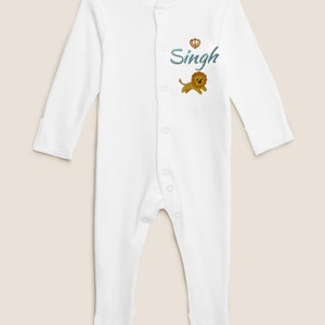 May include: White baby onesie with long sleeves and a snap closure. The onesie has an embroidered lion and the name "Singh" with a crown above it.