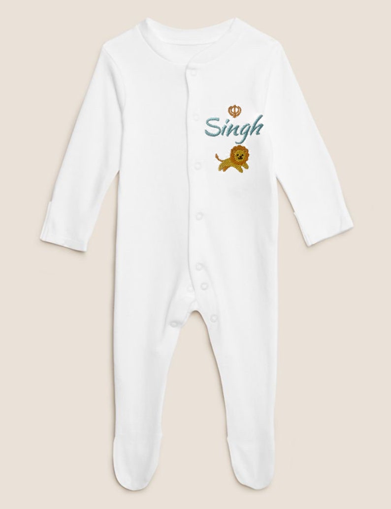 May include: White baby onesie with long sleeves and a snap closure. The onesie has an embroidered lion and the name "Singh" with a crown above it.
