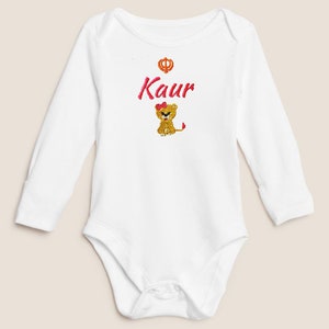 May include: White long-sleeved baby onesie with a pink embroidered lion and the word "Kaur" in pink lettering. A small gold symbol is above the word.
