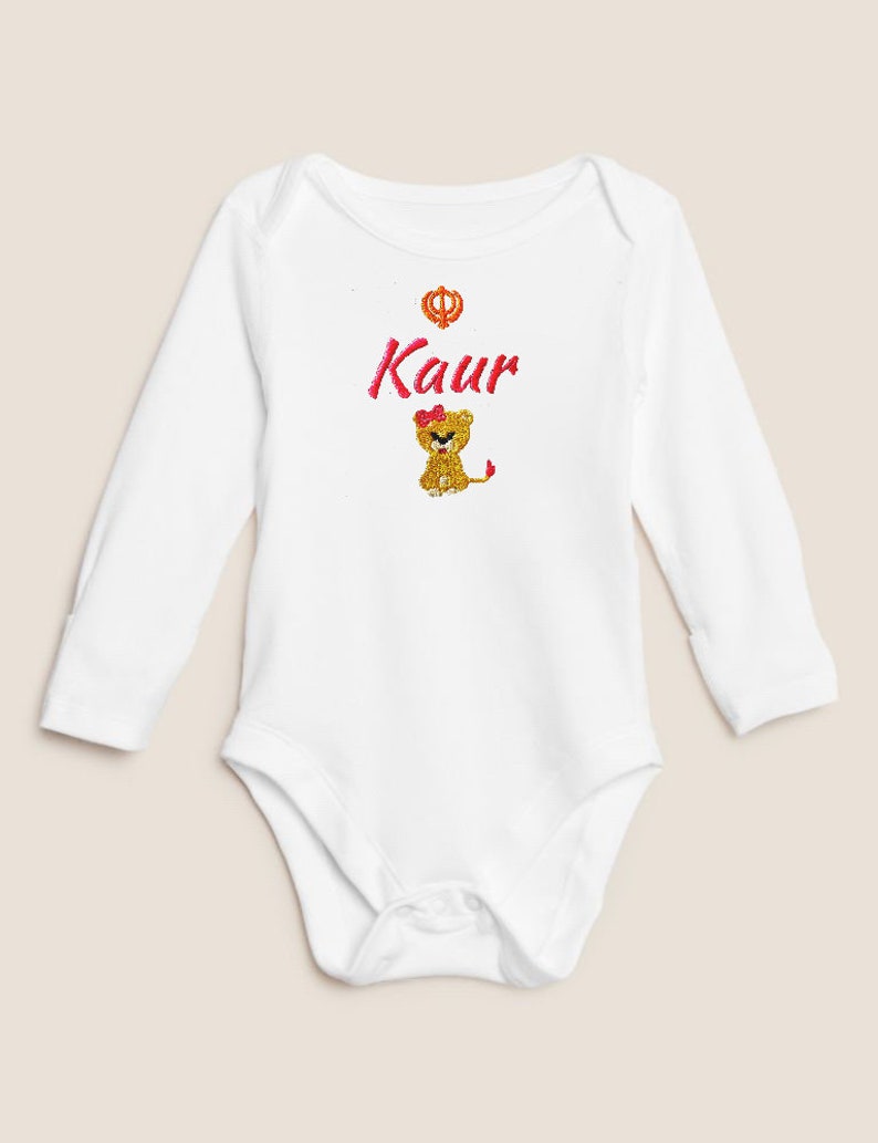 May include: White long-sleeved baby onesie with a pink embroidered lion and the word "Kaur" in pink lettering. A small gold symbol is above the word.
