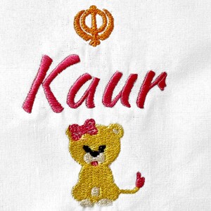 May include: Embroidered white fabric with the name "Kaur" in pink thread, a yellow lion with a pink bow, and a yellow and orange symbol.