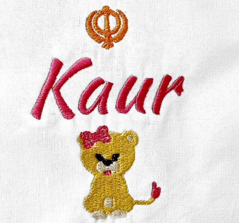 May include: Embroidered white fabric with the name "Kaur" in pink thread, a yellow lion with a pink bow, and a yellow and orange symbol.