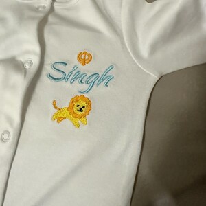 May include: White baby onesie with blue embroidered text "Singh" and a small yellow lion with a crown above the text.