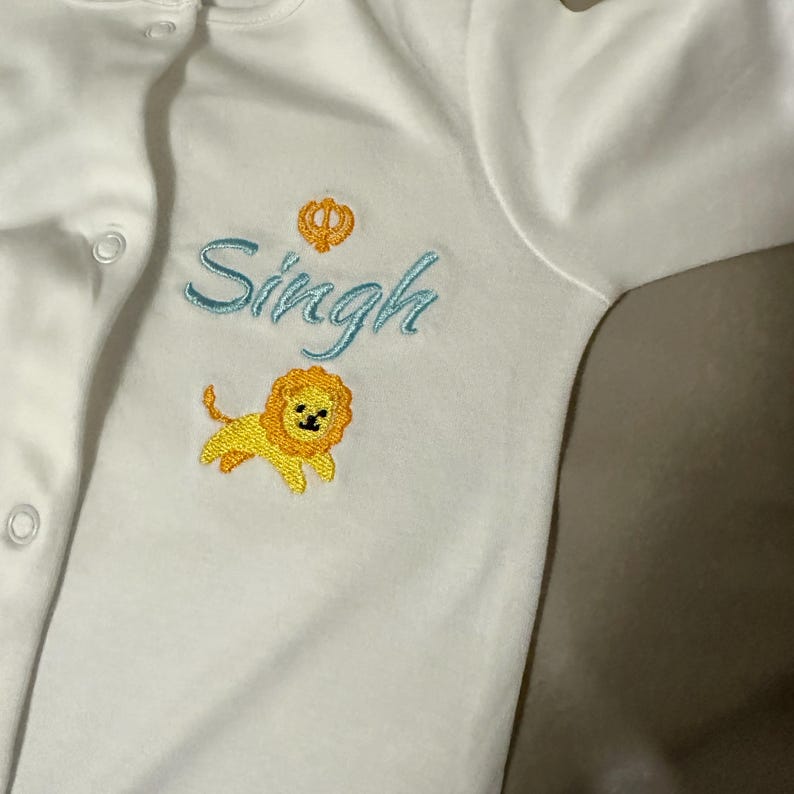 May include: White baby onesie with blue embroidered text "Singh" and a small yellow lion with a crown above the text.