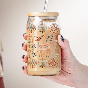 May include: A glass jar with a bamboo lid and a straw. The jar has a colorful design featuring a deer, trees, and geometric patterns. The jar is filled with a light brown liquid.