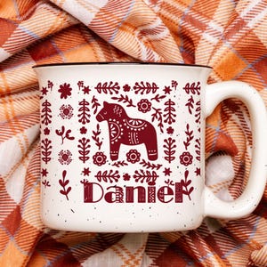 May include: White ceramic mug with a red and white Scandinavian folk art design featuring a horse and floral patterns. The mug has a black rim and handle and the name "Daniel" is printed on the front.