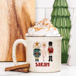 May include: A speckled ceramic mug filled with whipped cream dusted with cinnamon. The mug features illustrations of a mouse king, ballerina, and nutcracker, with the name "Sarah" in red. Cinnamon sticks rest in front of the mug.