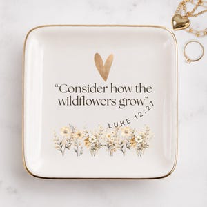 May include: A square white ceramic trinket dish with a gold-rimmed edge. The center features a gold heart icon above the black text "Consider how the wildflowers grow" and "LUKE 12:27," with a printed illustration of wildflowers at the bottom.