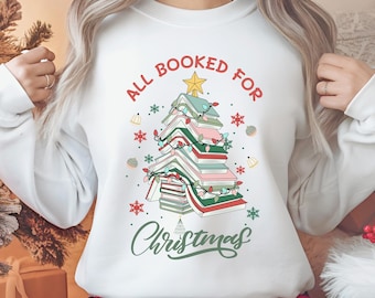 May include: White sweatshirt with a Christmas tree design made of books. The text "ALL BOOKED FOR CHRISTMAS" arches above the tree, with "Christmas" in a script font below. The tree is decorated with lights and a star.