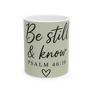 May include: A sage green ceramic mug featuring the black text Be still & know PSALM 46:10 written in a combination of cursive and serif fonts. A small black heart outline is positioned below the text on the front of the cylindrical mug.