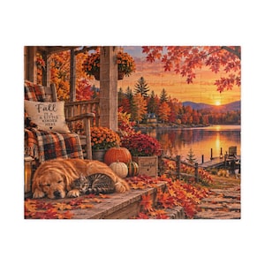 Pode incluir: Um quebra-cabeça retratando um golden retriever e um gato malhado descansando em uma varanda coberta de folhas de outono. Uma cadeira com uma almofada com a frase Fall is a little kinder here, abóboras e uma caixa com a palavra Thankful.